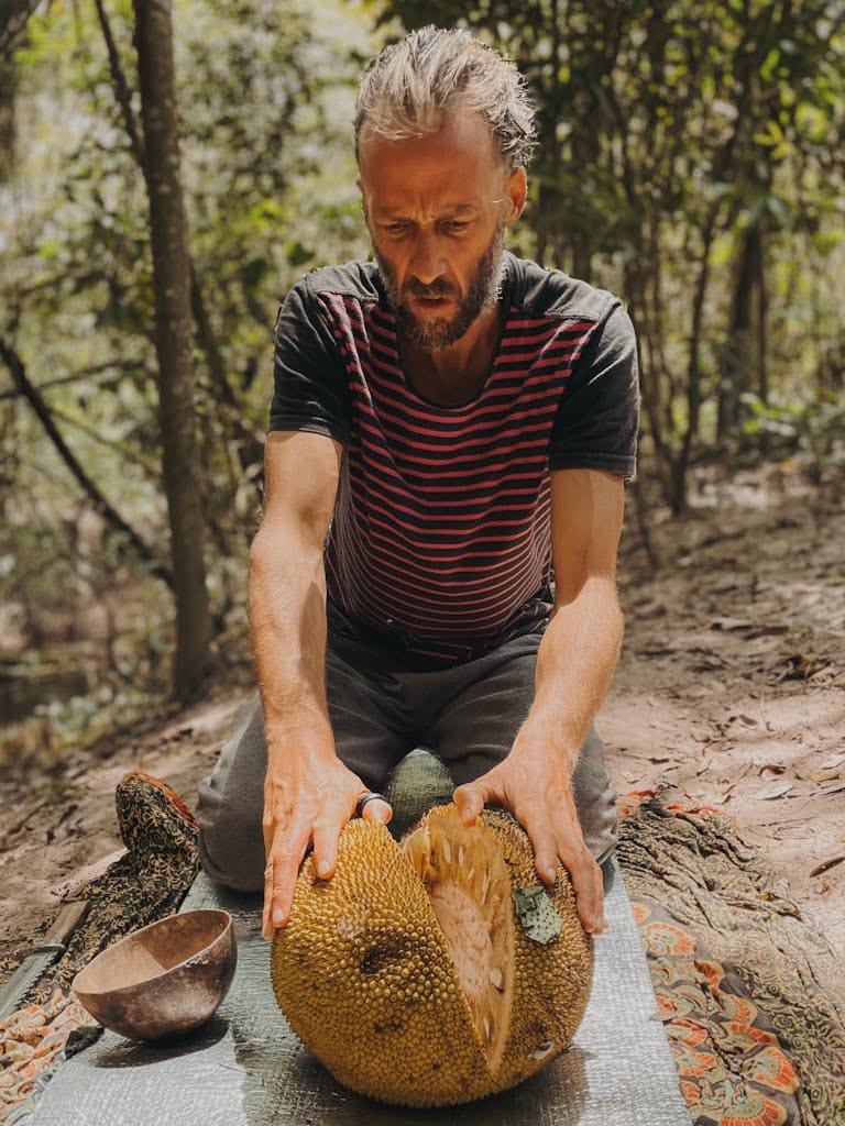 A man cutting a jackfruit outdoors in a forested area.