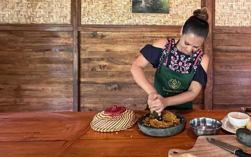 Chef demonstrating traditional Balinese cooking techniques by grinding spices with a cobek and ulekan in an open-air kitchen.