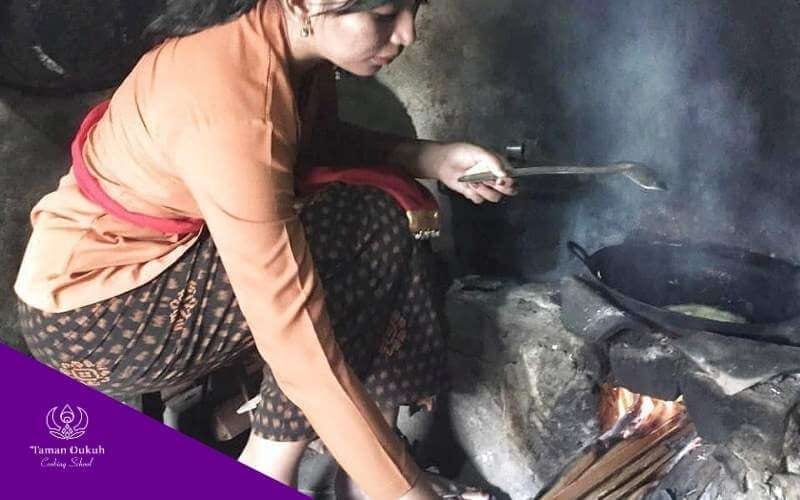A Balinese individual performing a symbolic purification ritual by warming hands at the bungut paon (mouth of the stove) to dispel negativity.