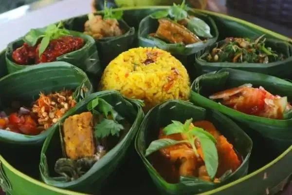 A traditional Balinese meal served on a large woven platter, featuring a central mound of yellow rice (Nasi Kuning) surrounded by various side dishes in individual banana leaf containers.